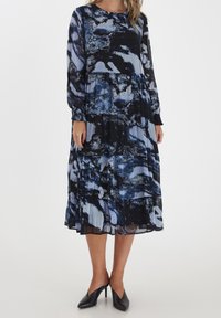 Vestido midi de manga larga en azul y negro con un patrón abstracto, hecho de una tela ligera con un diseño en capas y un ligero brillo. Tacones negros puntiagudos.