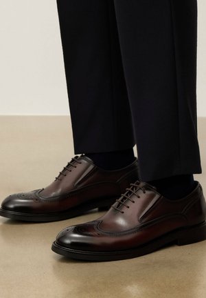 Brūnas ādas brogue kurpes ar melnām zolēm, valkātas ar tumšām biksēm uz bēša grīdas.