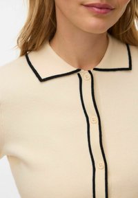 Kremowy sweter rozpinany na guziki z czarną lamówką, posiada kołnierz i zapięcie na pięć guzików. Gładka tekstura o klasycznym kroju.