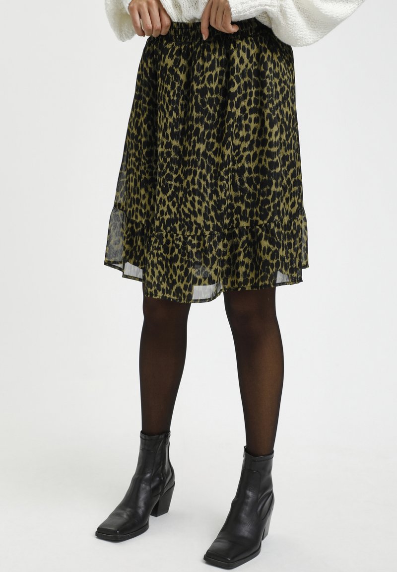 Femme portant une jupe imprimée léopard verte et noire, des collants noirs transparents et des bottines noires, debout sur un fond blanc.