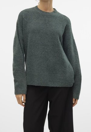 Frau trägt einen locker sitzenden dunkelgrauen Strickpullover mit langen Ärmeln und schwarze Hose, dargestellt von Hals bis Hüfte vor weißem Hintergrund.