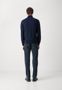 Pulover bleumarin cu guler înalt, manșete și tiv cu nervuri, asociat cu blugi închiși la culoare și pantofi negri. Materialul pare neted și ușor.