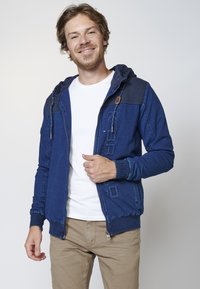 Blue zip-up hoodie with a textured shoulder panel, logo detail, and drawstring hood. Paired with a white t-shirt and khaki pants.