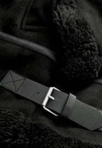 Zwarte nepbontjas met een gladde, gestructureerde oppervlakte, een brede kraag en een zwart leren riem met een zilveren gesp en stiksels.