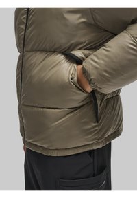 Olive roheline puhv jacket, millel on läikiv viimistlus, küljetasku ja mustad aktsendid servades ning tõmblukkudel.