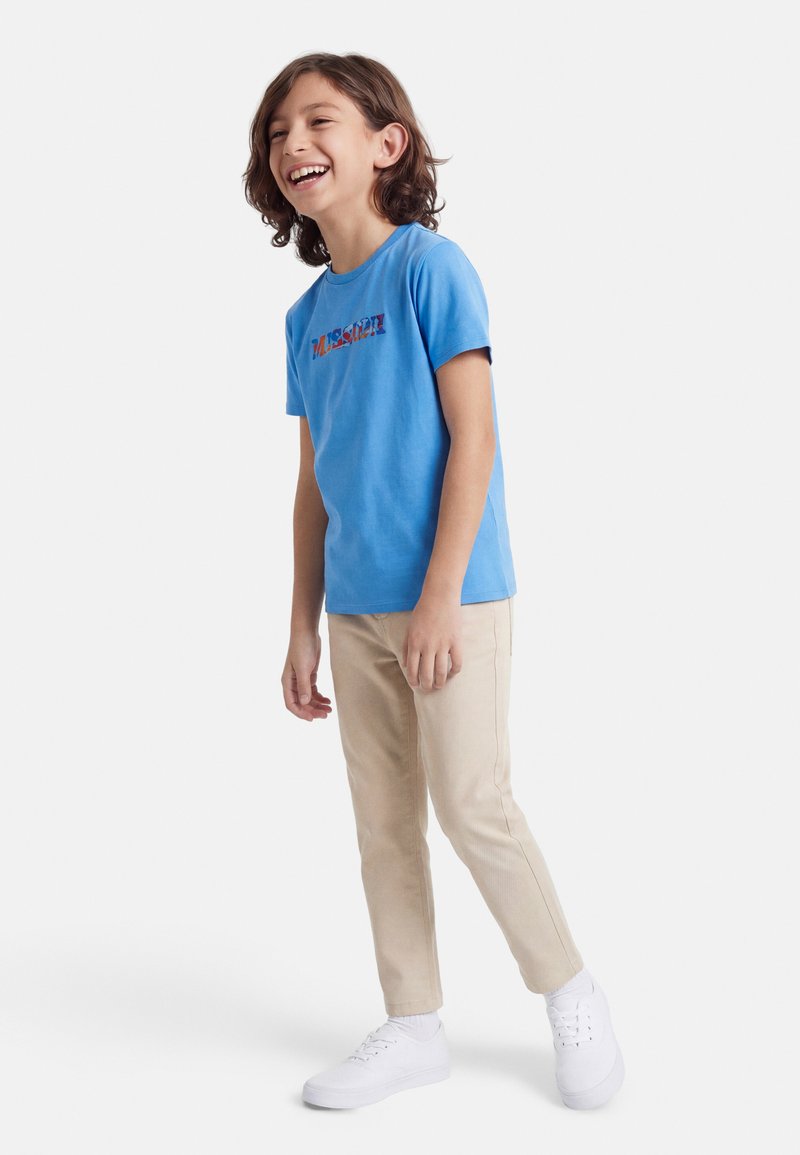 Αγόρι με μεσαίου μήκους καστανά μαλλιά που χαμογελά, φοράει μπλε μπλουζάκι, μπεζ παντελόνι, λευκές κάλτσες και λευκά αθλητικά παπούτσια, στέκεται σε λευκό φόντο.