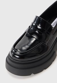 Svart lackläder loafer med en kraftig svart sula, prydd med en dekorativ rem, slät yta och rundad tådesign.