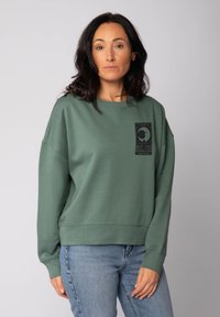 Sweatshirt vert à manches longues, coupe décontractée, ourlet côtelé, et un motif en noir de la lune sur le devant. Assorti avec un jean bleu clair.