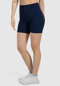 Pantaloni corti sportivi blu navy con una vita alta, realizzati in tessuto elastico. Texture liscia con cuciture piatte; abbinati a sneakers bianche.