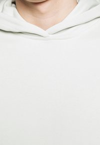 Sweat à capuche gris clair en tissu doux et texturé. Comprend une capuche avec cordon de serrage et une coupe décontractée, mettant en valeur un design simple et minimaliste.
