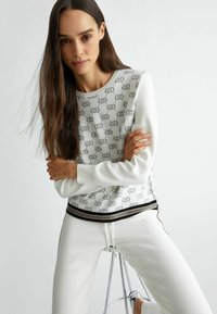 Maglione bianco con motivo decorativo, scollatura rotonda e dettagli in nero/oro all'orlo, abbinato a pantaloni bianchi. Tessuto morbido, vestibilità casual.