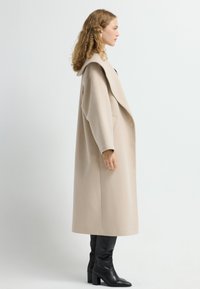 Beige uldfrakke med brede revers og sidelommer, parret med sort læderstøvler op til knæene med blokhæle.