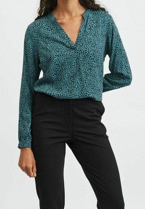 Vrouw draagt een teal gekleurde blouse met lange mouwen met zwarte onregelmatige vlekken en zwarte aansluitende broek tegen een effen achtergrond.