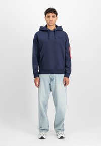 Ung mann står iført en marineblå hettegenser med lommer på forsiden og lyseblå, løse jeans med hvite joggesko.