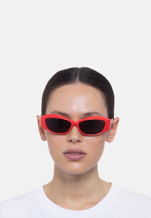 Lunettes de soleil rouges et angulaires avec des verres foncés et des montures en plastique. Comprend des branches élégantes avec des accents métalliques, portées sur une chemise blanche.