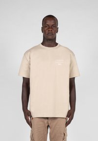 Beige cotton t-shirt with a crew neckline, short sleeves, and a small white graphic design on the front. Straight cut, soft texture.