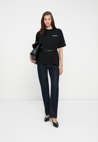 T-shirt nera oversize con testo bianco, cinto in vita, abbinato a jeans dritti scuri e ballerine nere. Tiene una clutch nera.