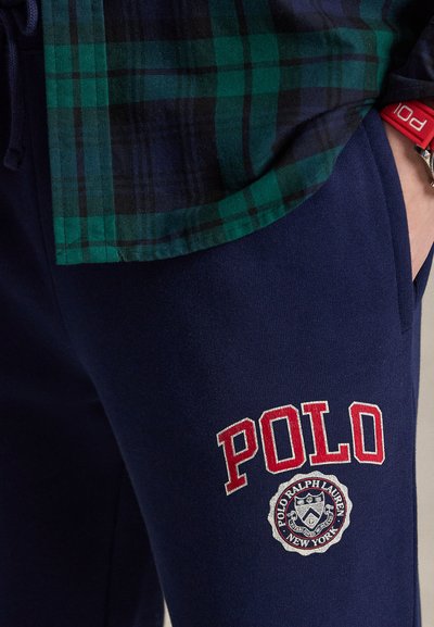 Σκούρα μπλε φόρμα με κόκκινο κείμενο "POLO" και λογότυπο Polo Ralph Lauren, συνδυασμένη με σκούρο πράσινο και σκούρο μπλε καρό πουκάμισο, χέρι στην τσέπη, κόκκινο βραχιόλι.