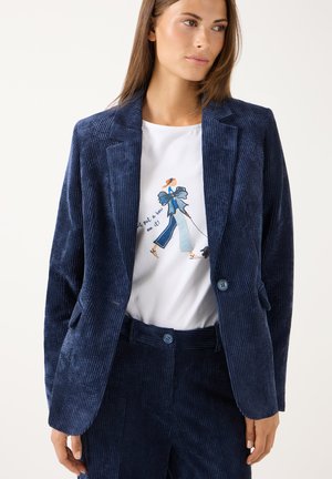 Vrouw die een blauwe corduroy blazer en broek draagt met een wit T-shirt met een afbeelding van een lopende vrouw en tekst.