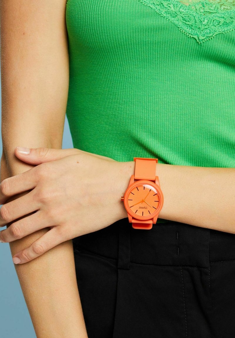 Oranges Silikonarmband mit rundem Zifferblatt und schwarzen Stundenmarkierungen, getragen am Handgelenk vor einem grünen Oberteil und schwarzem Hosenhintergrund.