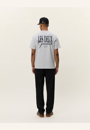Homme portant un t-shirt gris avec le texte « Les Deux Goods », un pantalon noir, des chaussures marron et une casquette noire, debout dos à la caméra sur un fond uni.