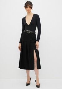 Vestido negro de manga larga con escote en V profundo, ceñido a la cintura con una hebilla dorada, con pliegues y una alta abertura lateral.