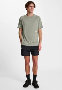 Maglietta sportiva a maniche corte verde chiaro, pantaloncini neri con tasche con zip, scarpe da ginnastica nere e calzini alla caviglia, in piedi contro uno sfondo uniforme.