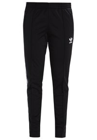 Svarta träningsbyxor med en elastisk midja, sidofickor med dragkedja och vita tre-rands detaljer längs sidorna. Har Adidas-logotyp.