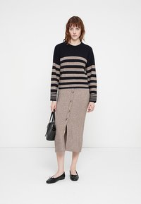 Woman wearing black and beige striped sweater, beige buttoned midi skirt, black flats, holding black handbag, standing against white background.