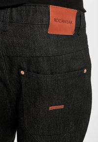 Black denim pants featuring a leather ROCAWEAR tag at the back waist and a side pocket with copper rivets. Textured fabric for durability.