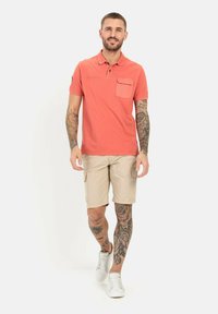 Koraalrode polo met een borstzak, gecombineerd met beige cargoshorts. Model draagt witte sneakers. Zichtbare tattoo's op armen en benen.
