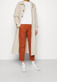 Trench-coat beige avec boutons marron, pull en tricot blanc, pantalon court orange et baskets blanches avec accent vert. Textures de tissu lisses.