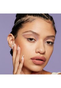 Gros plan sur un visage avec de longs ongles blancs pointus, des lèvres nude mates douces, un fard à paupières neutre avec une touche de violet et des cheveux nattés sur un fond lilas.