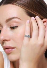 Zilveren ring met een golvend ontwerp, met een gladde oppervlakte en subtiele afgeronde randen, gedragen aan een hand dichtbij het gezicht.