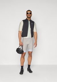 Tenue de cyclisme avec un gilet noir à fermeture éclair à l'avant et des accents bleu clair, un short gris clair, des chaussures de cyclisme noires et un casque noir.