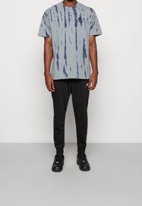 Šedé tričko s tie-dye vzorem, krátký rukáv a vyšité logo, v kombinaci s černými sportovními kalhotami a černými teniskami, stojící na neutrálním pozadí.