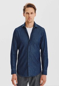 Granatowa koszula dżinsowa z zapięciem na guziki, długimi rękawami i ostrym kołnierzykiem. Noszona na białej koszulce, prezentująca swobodny, ale zarazem zorganizowany wygląd.