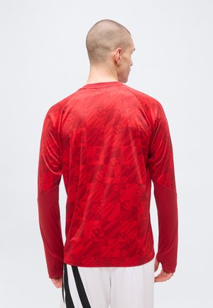 Homme à la tête rasée portant une chemise rouge à manches longues à motifs et un pantalon blanc avec des rayures noires, de dos sur un fond uni.