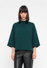 Maglia oversize verde scuro con collo alto e maniche corte e larghe, realizzata in un tessuto liscio, abbinata a pantaloni in pelle nera.