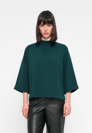Top azul oscuro de gran tamaño con cuello alto y mangas cortas y anchas, hecho de una tela suave, combinado con pantalones de cuero negro.