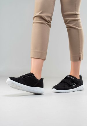 Osoba nosząca czarne sneakersy wsuwane z białymi podeszwami i beżowe krótkie spodnie, stojąca na jednolitym jasnoszarym tle.