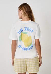 Biała bawełniana koszulka z nadrukiem żółtych cytryn i zielonych liści, z napisem "UN ZEST D'AMOUR" w niebieskim kolorze, luźny krój.
