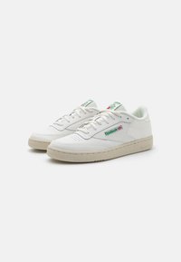 Sneakers in pelle bianca con punta perforata, fodera morbida, suola in gomma beige, marchio verde e rosso sulla linguetta e sul lato, design classico a bassa silhouette.