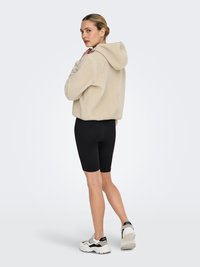Beige fleece hoodie with a textured surface, featuring a hood and cropped design, paired with black cycling shorts and white sneakers.