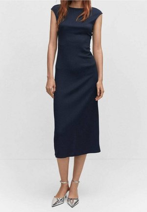 Kobieta ubrana w granatową sukienkę midi bez rękawów, w połączeniu z srebrnymi, spiczastymi butami na obcasie z paskiem wokół kostki, stoi na tle jednolitego tła.