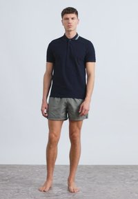 Tamsiai mėlynas polo marškinėliai trumpomis rankovėmis su kontrastine apykakle. Derinami su pilkais ir baltais dryžuotais šortais. Basas ant tekstūruoto paviršiaus.