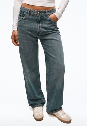 Osoba nosiaca modré džínsy s vysokým pásom a širokými nohavicami, biely dlhý crop top a biele tenisky, jedna ruka v vrecku.