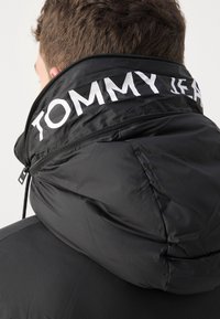 Schwarze Puffjacke mit hohem Kragen, im weißen Druck mit "TOMMY JEANS"-Logo, mit Reißverschluss und strukturiertem Stoff.
