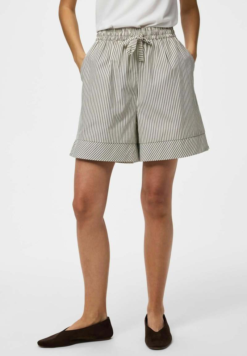 Femme portant un short beige et noir à fines rayures, longueur genoux, avec taille à cordon et ourlets retroussés, associé à des chaussures plates marron foncé.