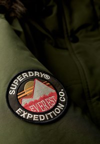 Tkanina oliwkowego parka posiada okrągłą czarną łatę z kolorowym wzorem gór oraz haft "SUPERDRY® EXPEDITION CO.".